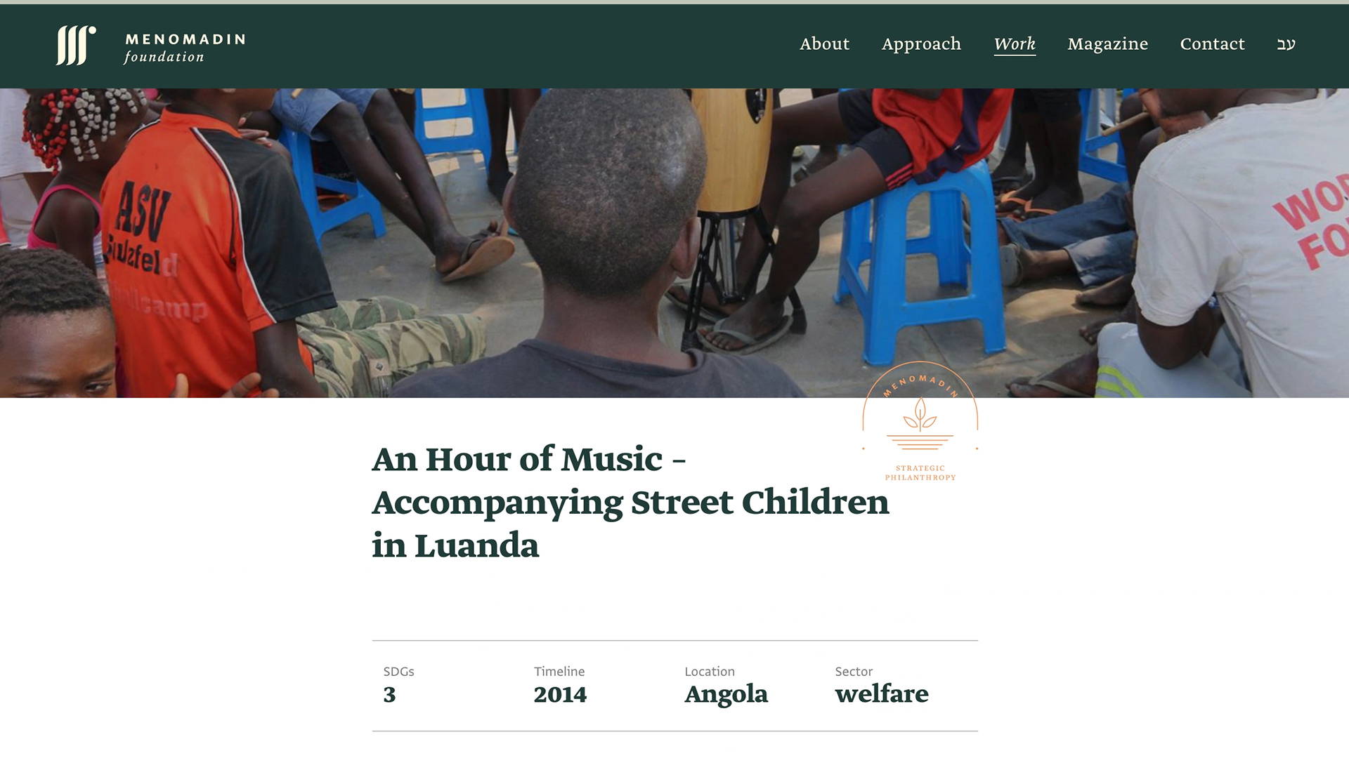The image shows a webpage from the Menomadin Foundation website featuring a header that reads “An Hour of Music - Accompanying Street Children in Luanda.” Below the header is a photograph showing the back of a person wearing a blue shirt, facing a group of children sitting on blue plastic chairs. The children are focused on something in front of them, not visible in the image. The webpage includes icons indicating three SDGs (Sustainable Development Goals), with a timeline marked as 2014, location as Angola, and sector as welfare.